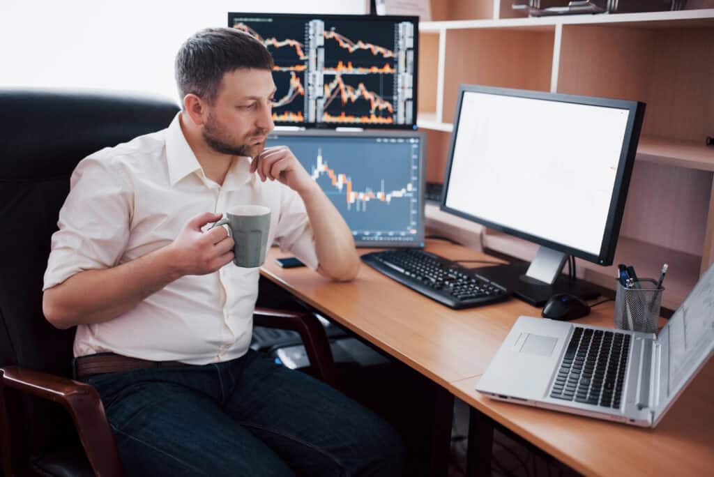 young businessman is sitting office table working computer with many monitors diagrams monitor stock broker analyzes binary options charts hipster man drinking coffee studying